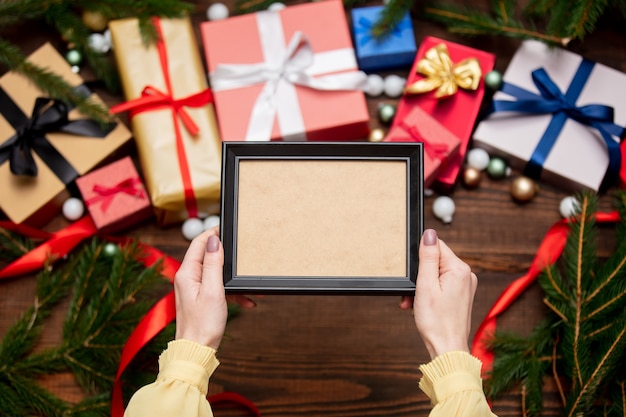 Mão feminina segurando porta-retrato perto com presentes de Natal e galhos de pinheiro com enfeites na mesa de madeira