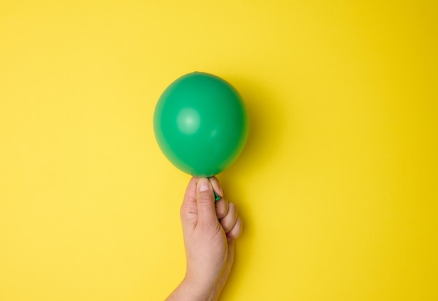 Mão feminina segurando um balão de ar verde inflado em um fundo amarelo, close-up