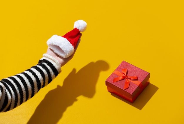 Mão feminina segurando um chapéu de Papai Noel perto de uma caixa de presente na superfície amarela