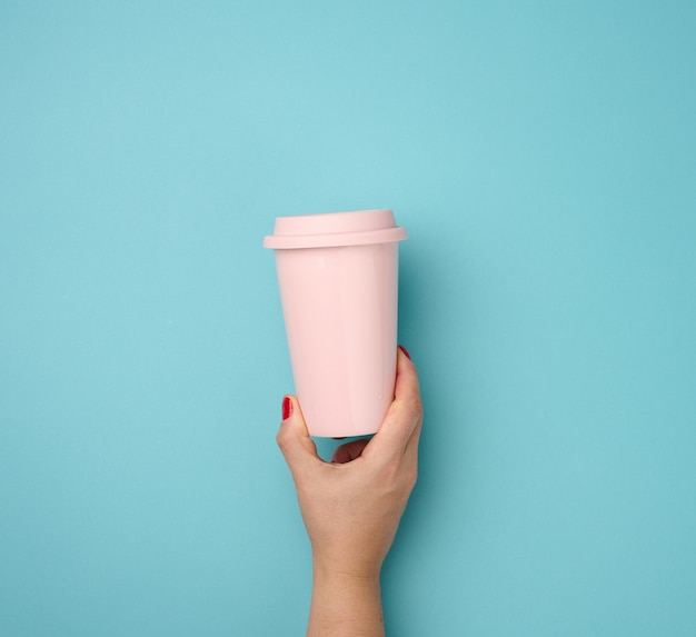 Mão feminina segurando uma caneca de cerâmica rosa com tampa de plástico em um fundo azul, intervalo e beber café