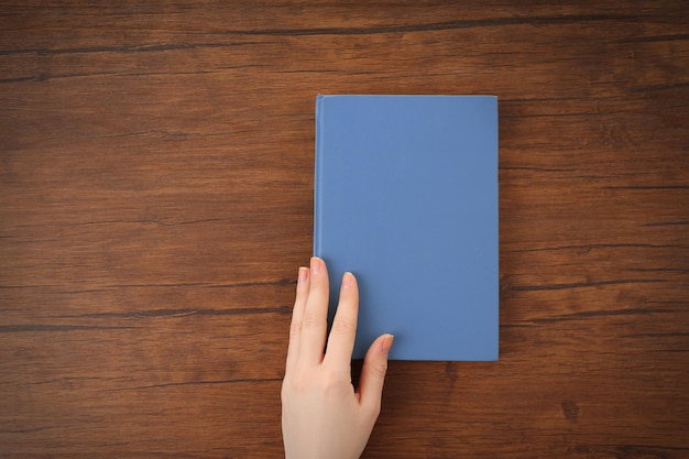 Mão feminina segurando uma capa de livro azul claro na vista superior da mesa de madeira marrom