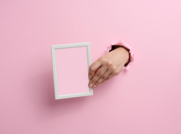 Mão feminina segurando uma moldura de madeira branca vazia, parte do corpo saindo de um buraco rasgado no fundo de papel rosa