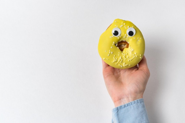 Mão feminina segurando uma rosquinha amarela engraçada com olhos enormes. Comida com caretas em fundo branco.