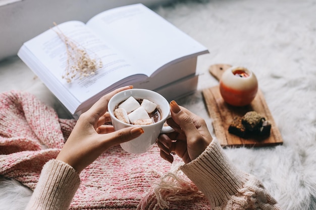 Mão feminina segurando uma xícara de marshmallow de chocolate quente e livro na temporada de inverno