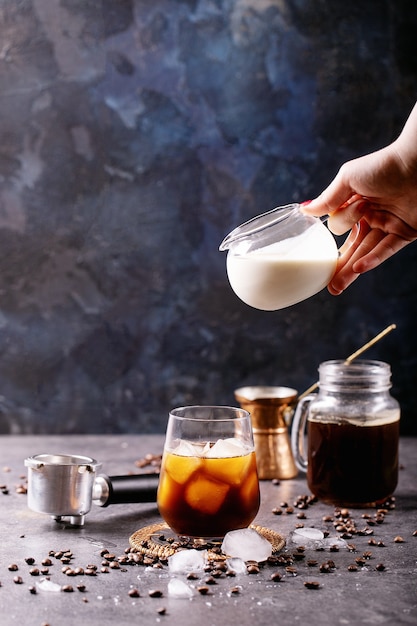 Mão feminina servindo creme no café gelado sobre a parede azul com grãos de café e cubos de gelo ao redor