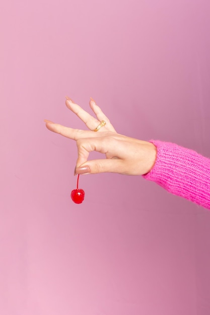 Mão feminina vestindo suéter rosa e segurando uma cereja. Fundo rosa claro.