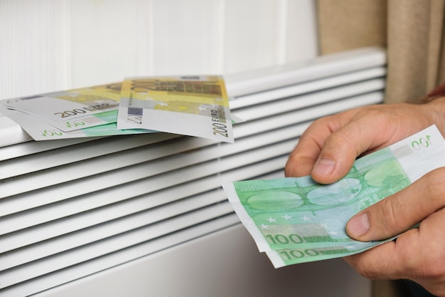 Mão humana segurando notas de euro perto de radiador de aquecedor elétrico em casa. Homem paga dinheiro para aquecimento.