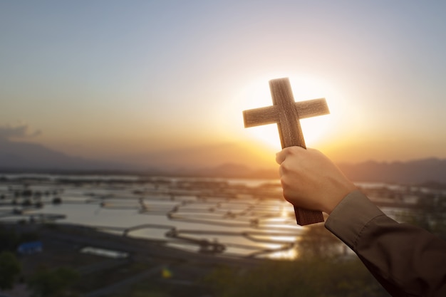 Mão humana segurando uma cruz cristã com um fundo do céu ao nascer do sol