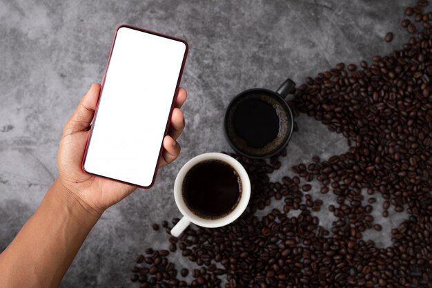 Mão humana segurar a tela em branco no telefone inteligente, telefone celular, tablet na xícara de café preto e grãos de café.