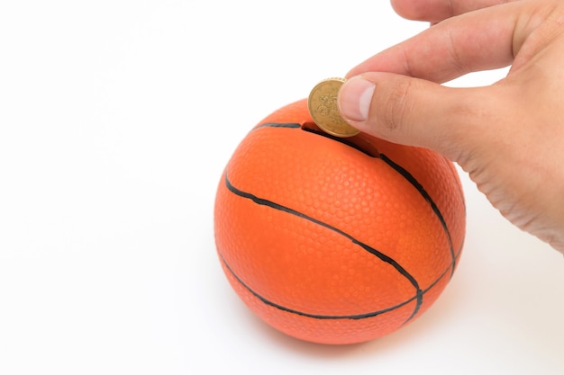 Mão masculina colocando moeda de euro no cofrinho com uma forma de bola de basquete isolada no fundo branco