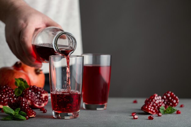 Mão masculina derramando suco de romã de uma garrafa em um copo. nas proximidades existem várias romãs.