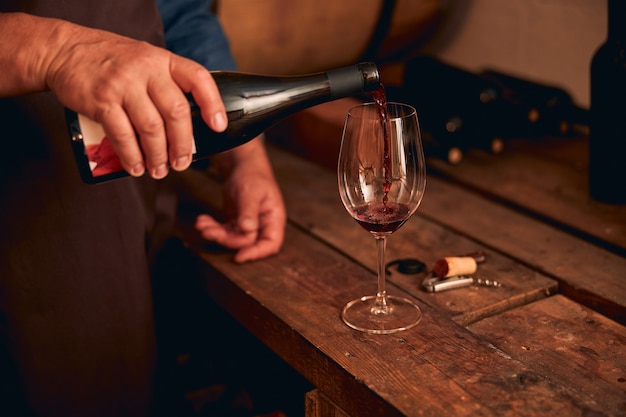 mão masculina despejando bebida alcoólica em um copo de vinho enquanto um homem em pé junto à mesa de madeira na adega