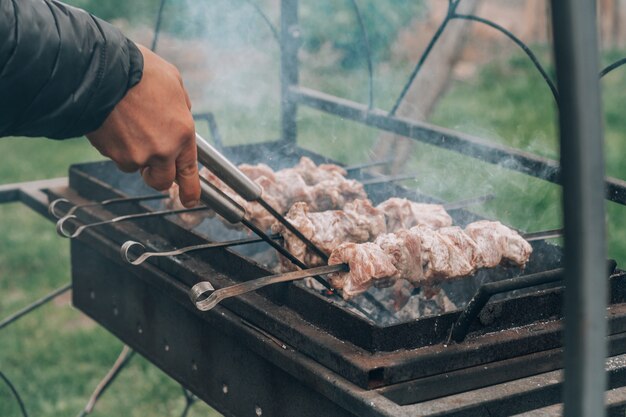 Mão masculina gira com pinças de carne crua frita em espetos, cozidos na grelha a carvão. Cozinhar churrasco ao ar livre