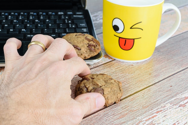 Mão masculina pegando um biscoito com gotas de chocolates. caneca amarela com leite e um laptop. Pausa para e-learning.