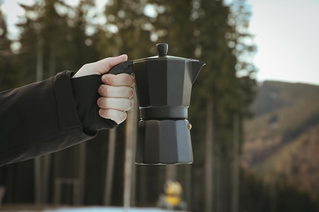 Mão masculina segura a cafeteira contra floresta e montanhas