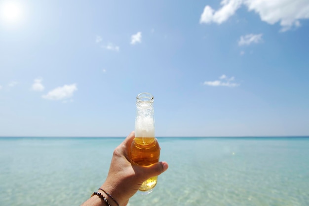 Mão masculina segurando uma garrafa de cerveja contra um céu ensolarado e mar cristalino Conceito de férias