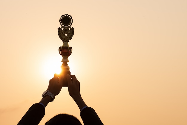 Mão mostrando o troféu de ouro premiado durante o pôr do sol. Conceito de sucesso do negócio.