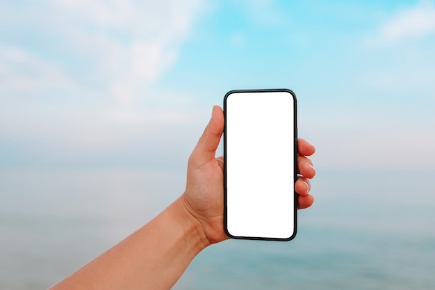 Mão mostrando um telefone inteligente em branco na praia com o mar ao fundo. Simulação de tela branca