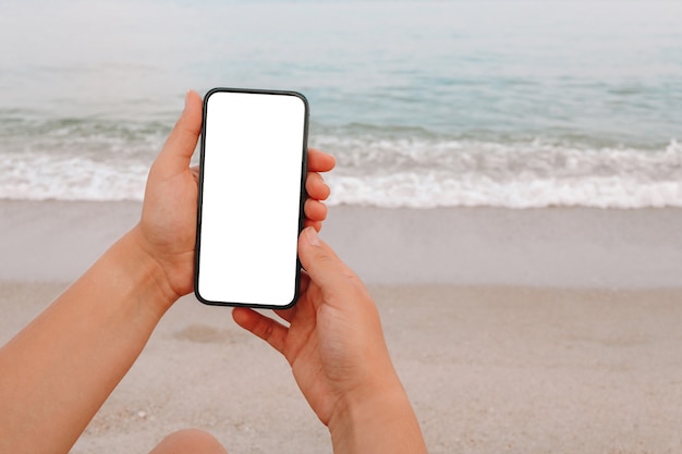 Mão mostrando um telefone inteligente em branco na praia com o mar ao fundo. simulação de tela branca