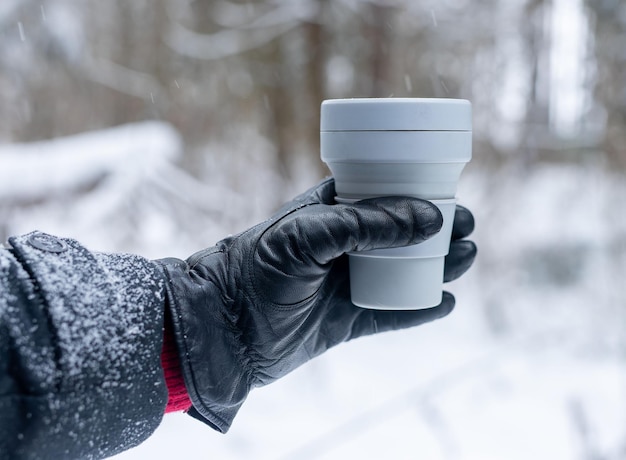 Mão na luva, segurando a xícara de café de silicone ecológico para viagem, tire a caneca no inverno em tempo de neve