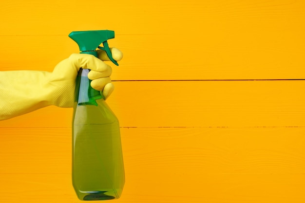 Mão na luva segurando spray de detergente doméstico contra um fundo amarelo de madeira