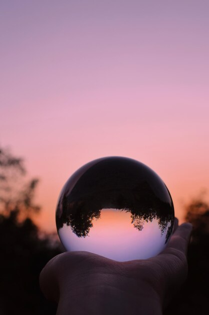 Foto mão recortada de pessoa segurando uma bola de cristal contra o céu durante o pôr do sol