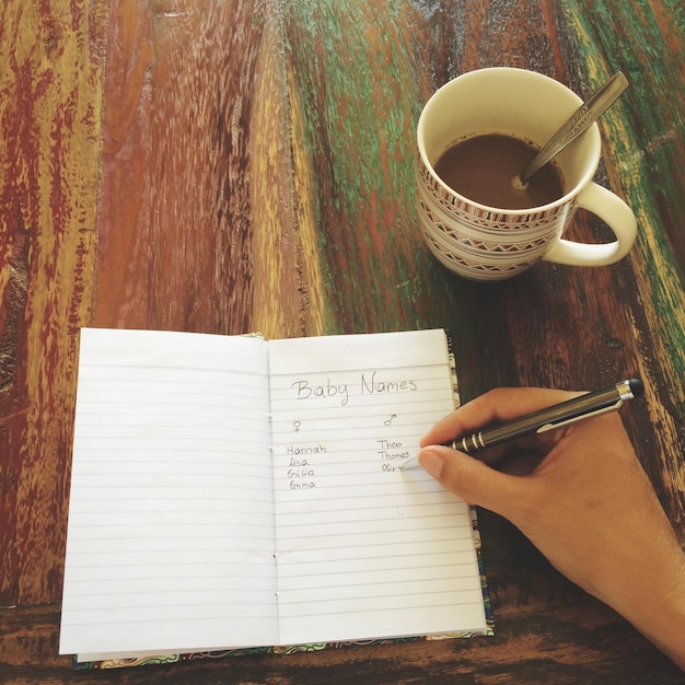 Foto mão recortada planejando nomes de bebês em caderno com café na mesa