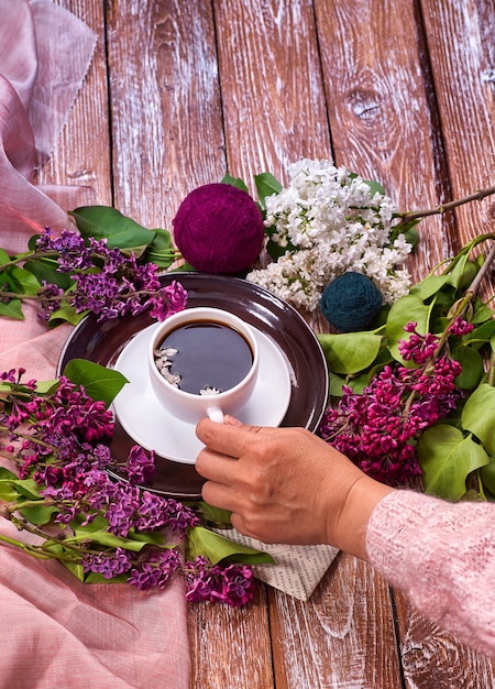 Mão segura uma xícara de café da manhã com ramos de flores lilás primavera desabrochando na vista de fundo de madeira de cima.