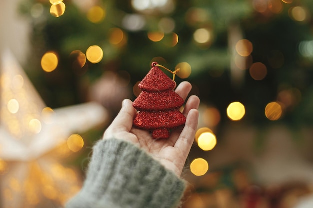 Mão segurando a árvore de natal vermelha no fundo da árvore iluminada com luzes douradas Mulher decorando a árvore de natal e segurando o enfeite de glitter na mão na sala festiva Tempo atmosférico