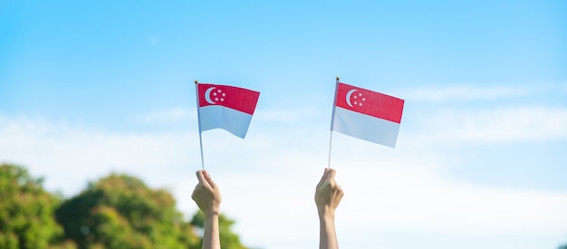 Mão segurando a bandeira de singapura no fundo do céu azul. dia nacional de cingapura e conceitos de celebração feliz