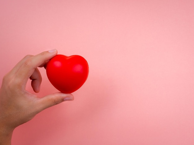Mão segurando a bola de coração vermelho sobre fundo rosa pastel com espaço de cópia. Conceito de amor, carinho, generosidade, bem-estar e conceitos de doação, estilo minimalista.