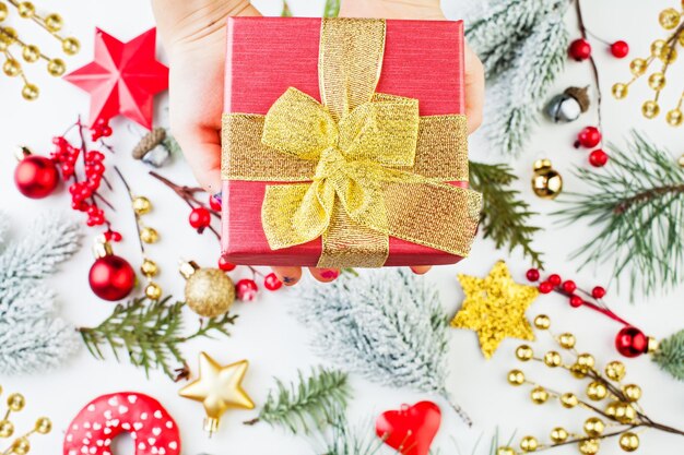 Mão segurando a caixa de presente vermelha com fita dourada e arco contra fundo de decoração de Natal Composição de Natal plana vista de cima