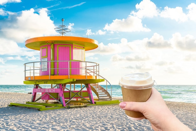 Mão segurando a xícara de papel de café quente e cabine ao nascer do sol em Miami Beach, Flórida, EUA