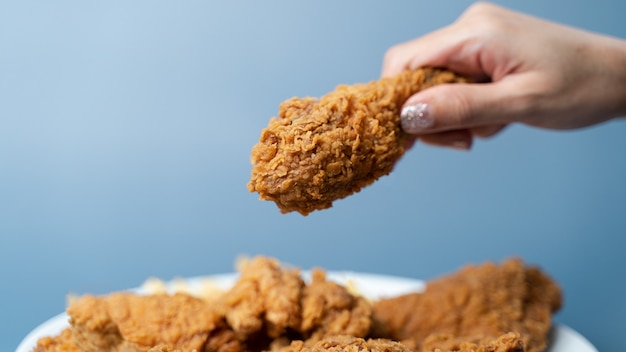 Mão segurando baquetas, frango frito crocante em chapa branca sobre fundo azul.