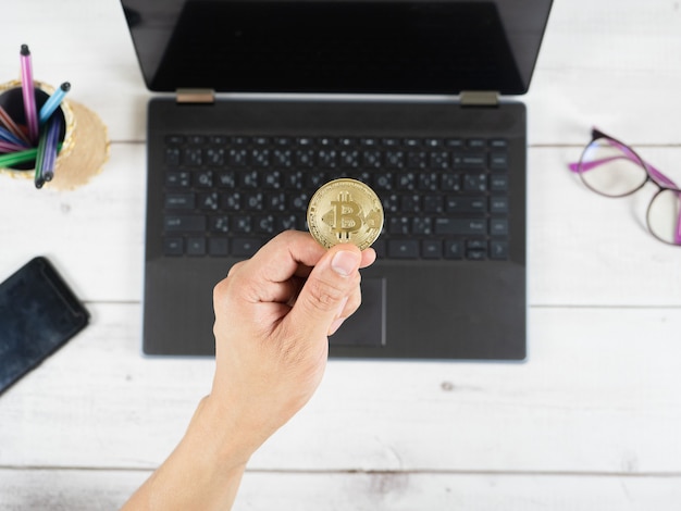 Mão segurando bitcoin no teclado do plano de fundo do laptop na área de trabalho da vista de cima da mesa