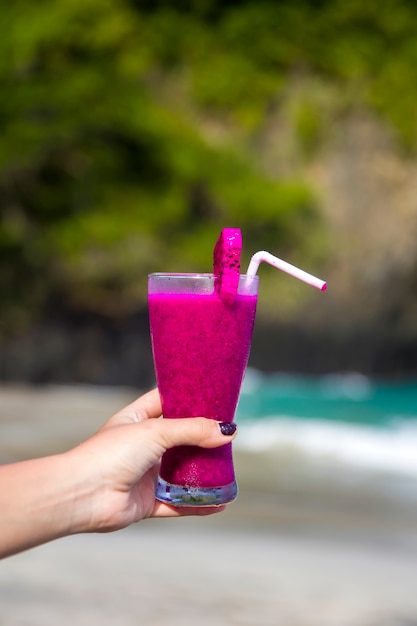 Mão segurando cocktail roxo em vidro decorado com canudos de plástico e pedaço de fruta do dragão na praia tropical