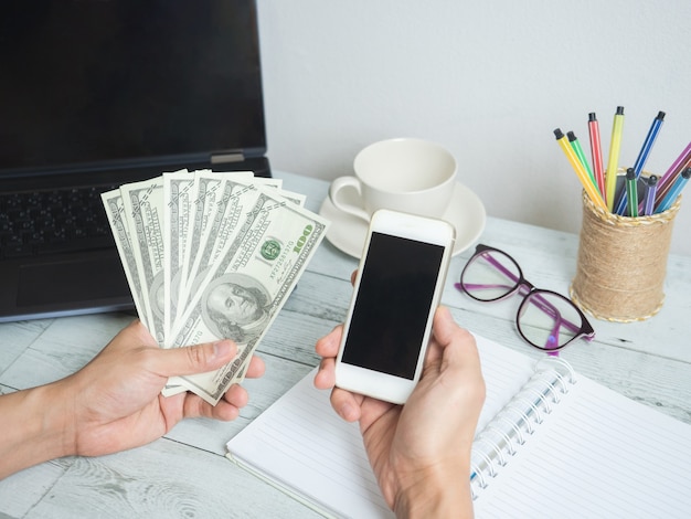 Mão segurando dinheiro, dólar e celular com área de trabalho no fundo da mesa de madeira branca