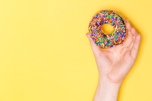 Mão segurando donut de chocolate fosco com granulado, glaceado com açúcar glaceado em fundo amarelo.