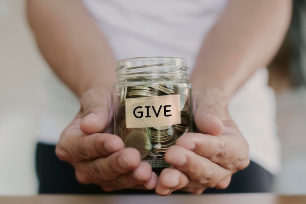 Mão segurando moedas em frasco de vidro com rótulo de texto escrito palavra GIVE para dar e conceito de doação