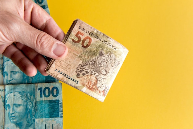 Mão segurando notas de dinheiro real brasileiro em fundo amarelo 50 notas reais