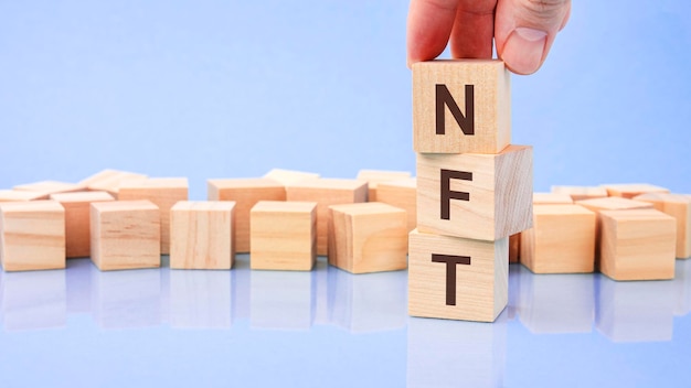 Mão segurando o bloco de cubo de madeira com texto NFT, a inscrição nos cubos é refletida no fundo azul da superfície com espaço de cópia NFT curto para token não fungível