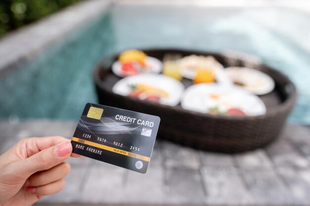 Mão segurando o cartão de crédito com café da manhã flutuante na piscina em viagens de luxo de pagamento de villa de luxo