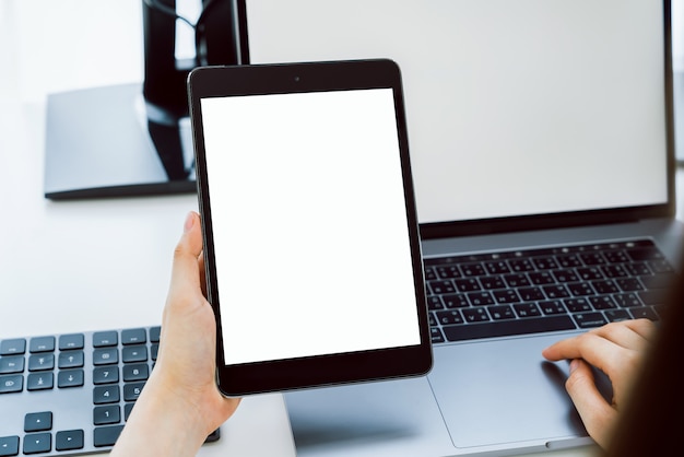 Mão segurando o modelo digital tablet de tela em branco e computador no escritório de mesa.