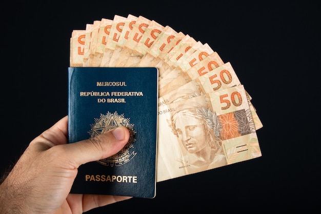 Mão segurando o passaporte brasileiro com notas de cinquenta reais dinheiro brasileiro