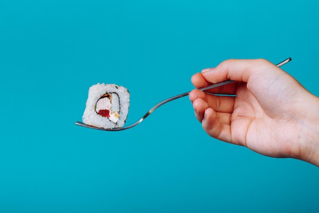 Mão segurando o rolo de sushi fresco com um garfo, isolado sobre fundo azul. Como comer sushi sem pauzinhos