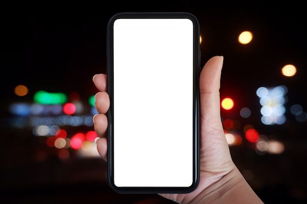 Mão segurando o smartphone com tela em branco sobre semáforo turva à noite