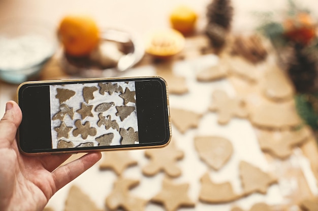 Foto mão segurando o telefone e tirando foto do processo de fazer biscoitos de gengibre de natal tirando foto no smartphone de biscoitos de gengibre crus na mesa rústica