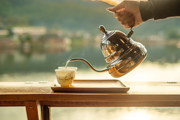 Mão segurando um bule vintage e servindo chá quente na xícara na mesa de madeira com vista para o lago