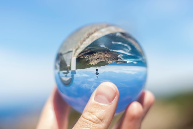 Mão segurando uma bola de cristal refletindo o belo e turístico farol da cidade galega de Ribadeo em frente ao mar, durante um dia ensolarado, conceitos, foco seletivo.