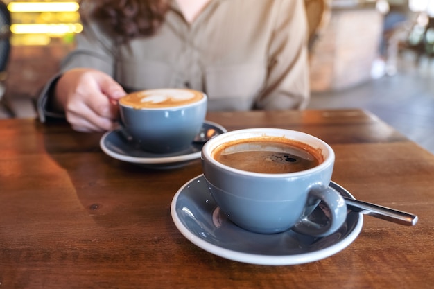 mão segurando uma xícara azul de café preto quente com outra xícara de café com leite na mesa de madeira no café